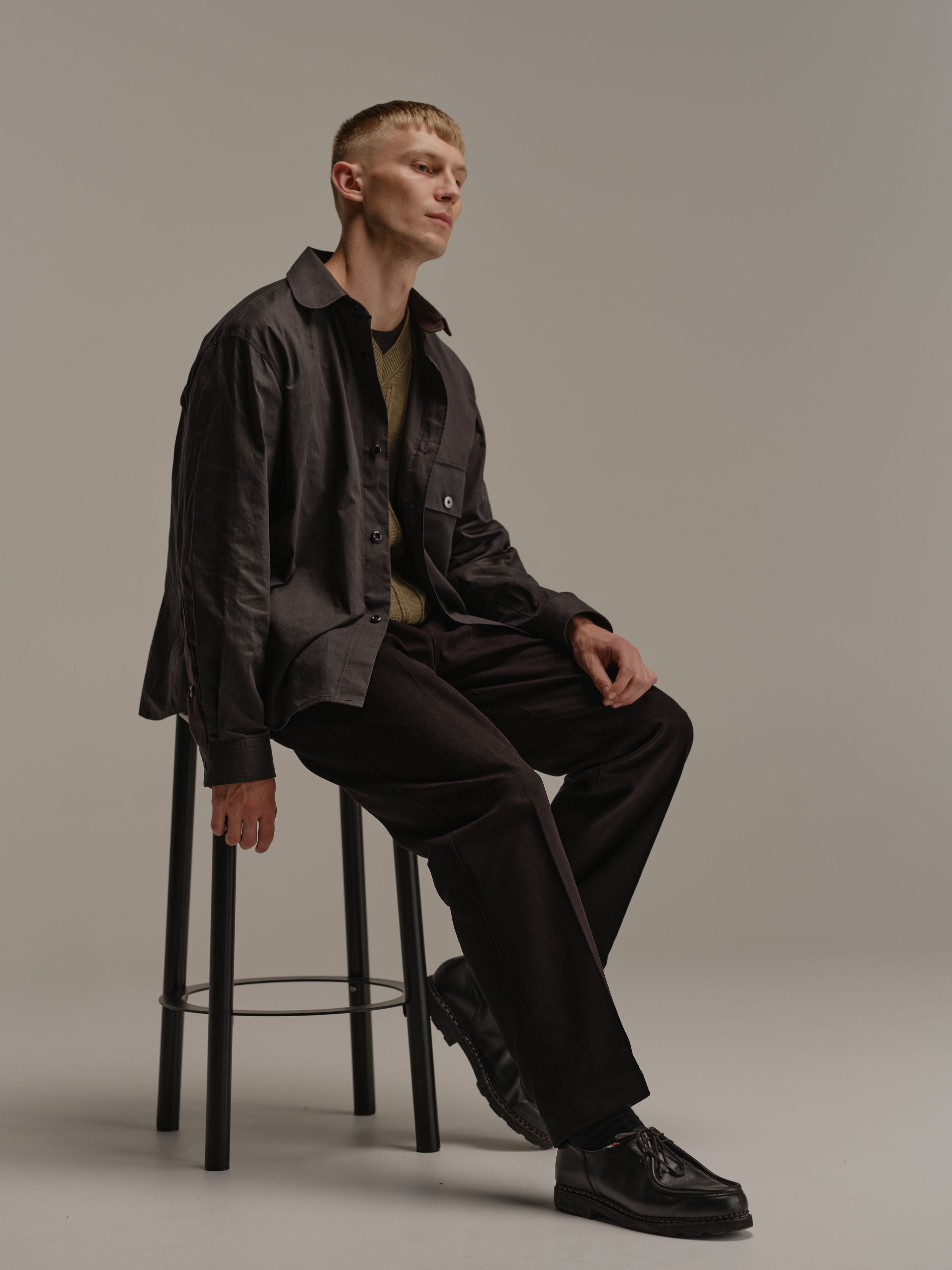 Man looks to his left while sitting on black metal stool wearing a black shirt, sage vest, black pants and black shoes.
