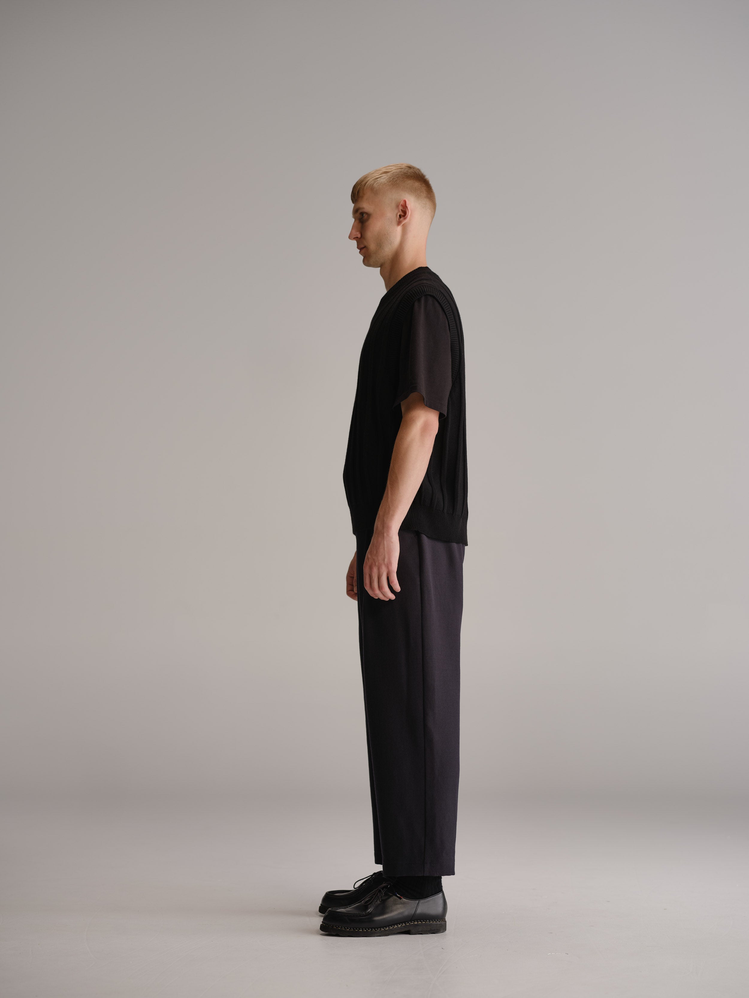 Side view of man in white studio wearing black vest over black t-shirt, navy wool trousers and black leather shoes.