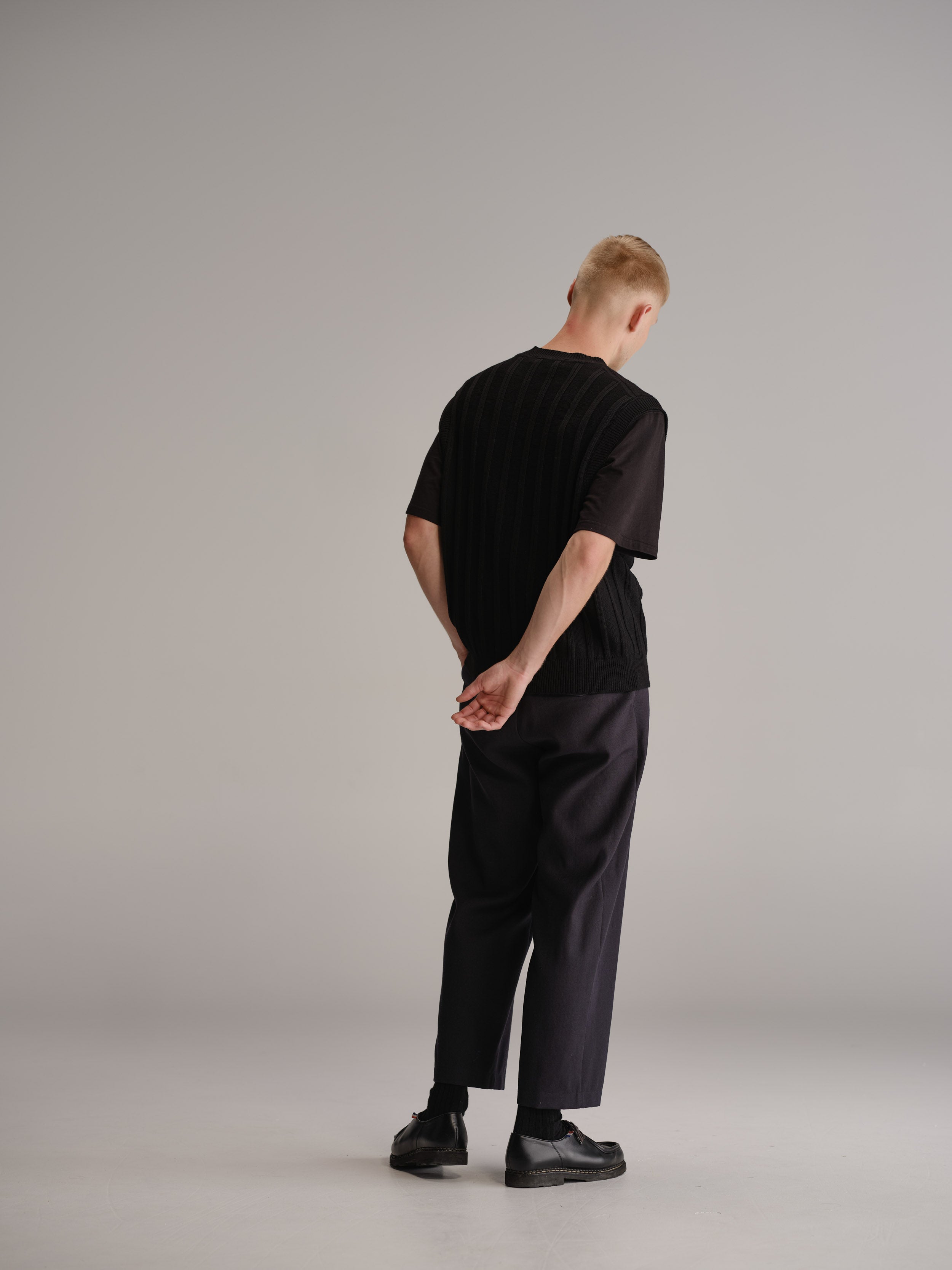 Back view of man in white studio wearing black vest over black t-shirt, navy wool trousers and black leather shoes.