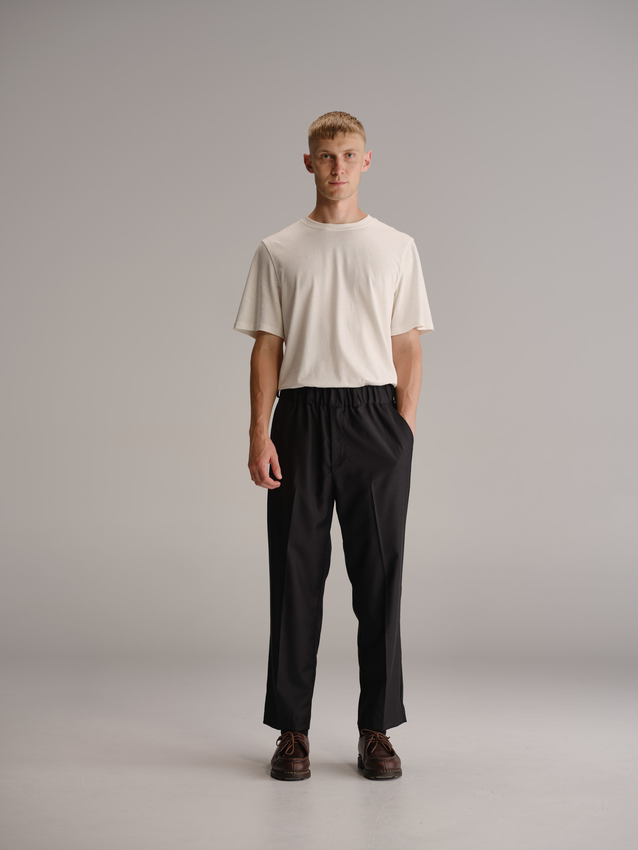 Front view of man standing in white studio wearing a tucked in white t-shirt and black trousers with brown leather shoes.