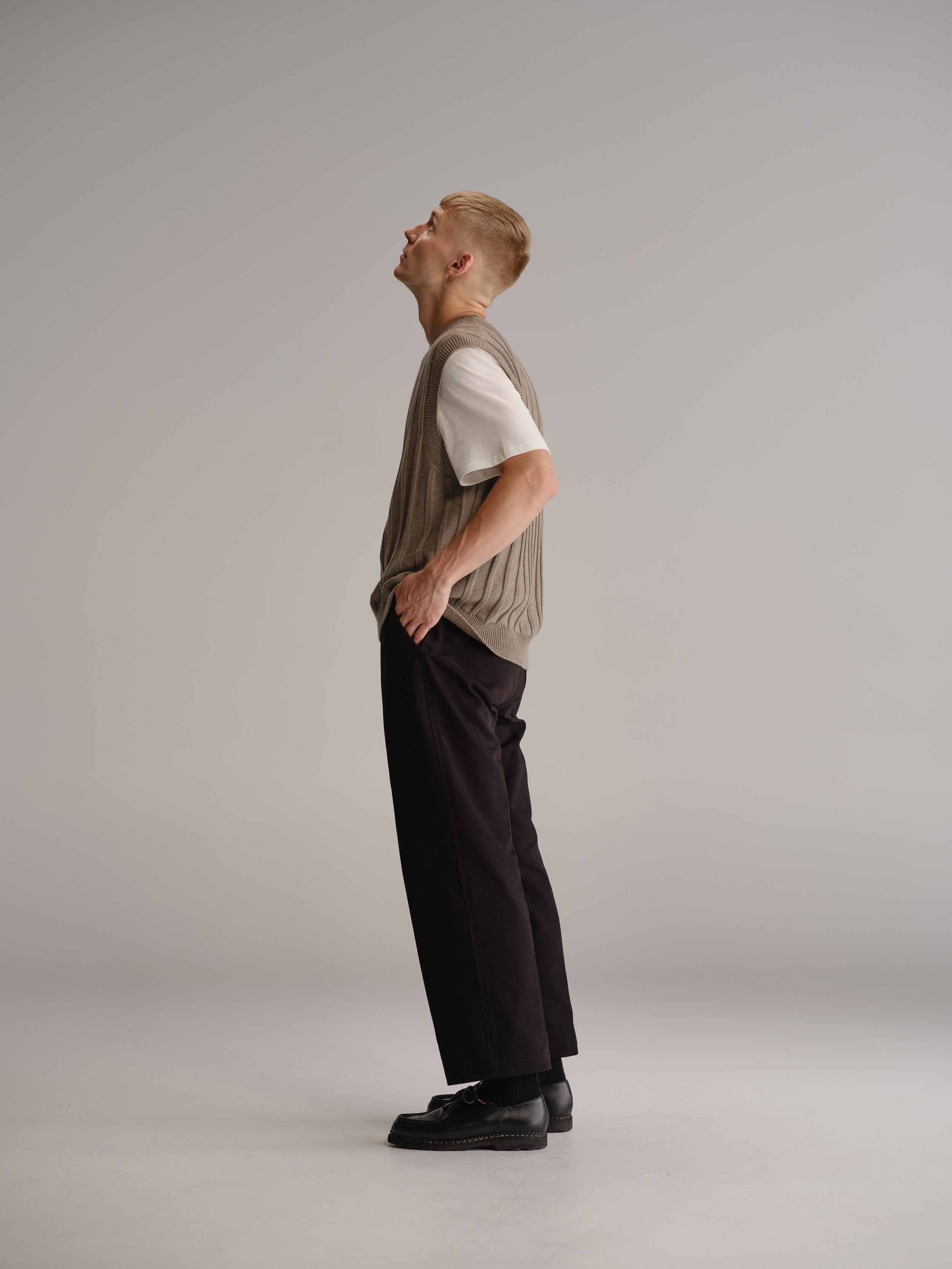 Side view of man standing in white studio wearing wheat vest over a white t-shirt, black pants and black leather shoes.
