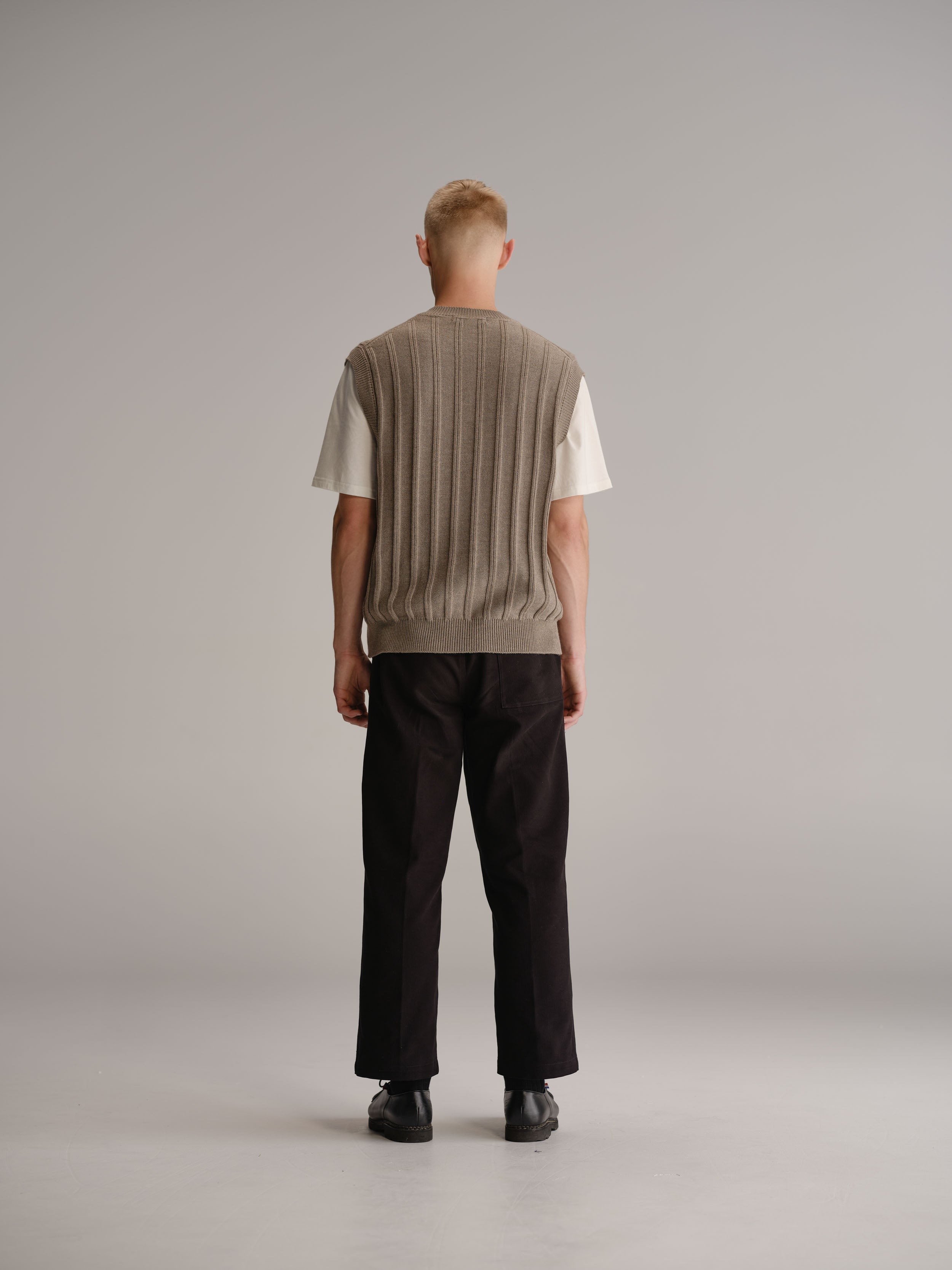 Back view of man standing in white studio wearing wheat vest over a white t-shirt, black pants and black leather shoes.