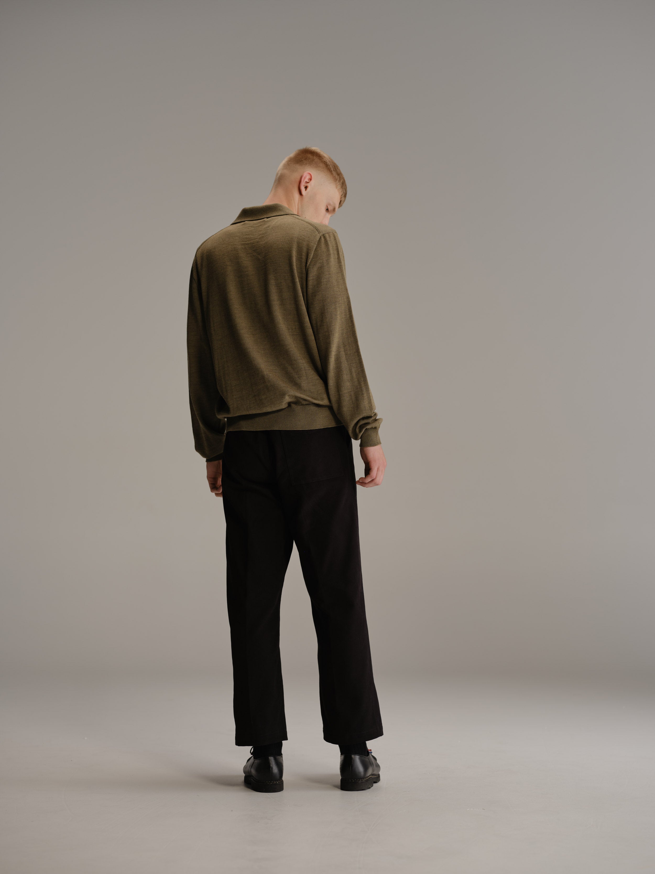 Back view of man standing in white studio looking to his right wearing sage polo shirt, black pants and black leather shoes.