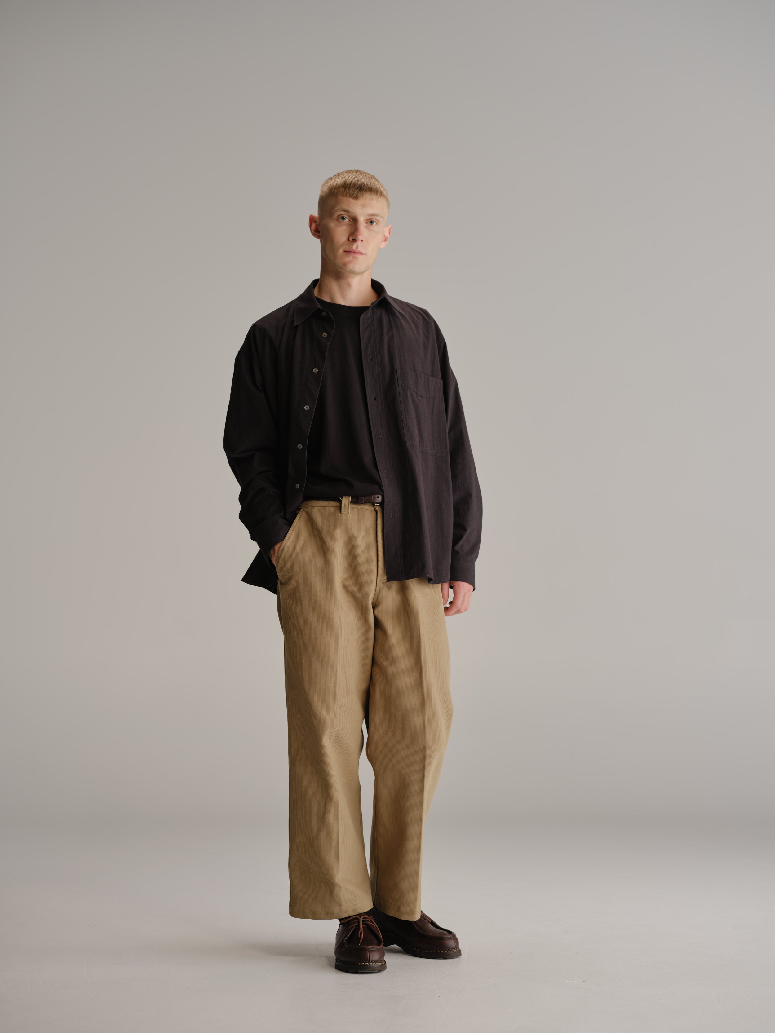 Front view of man standing in white studio with hand in pocket wearing black shirt, tucked in black t-shirt and fawn pant.
