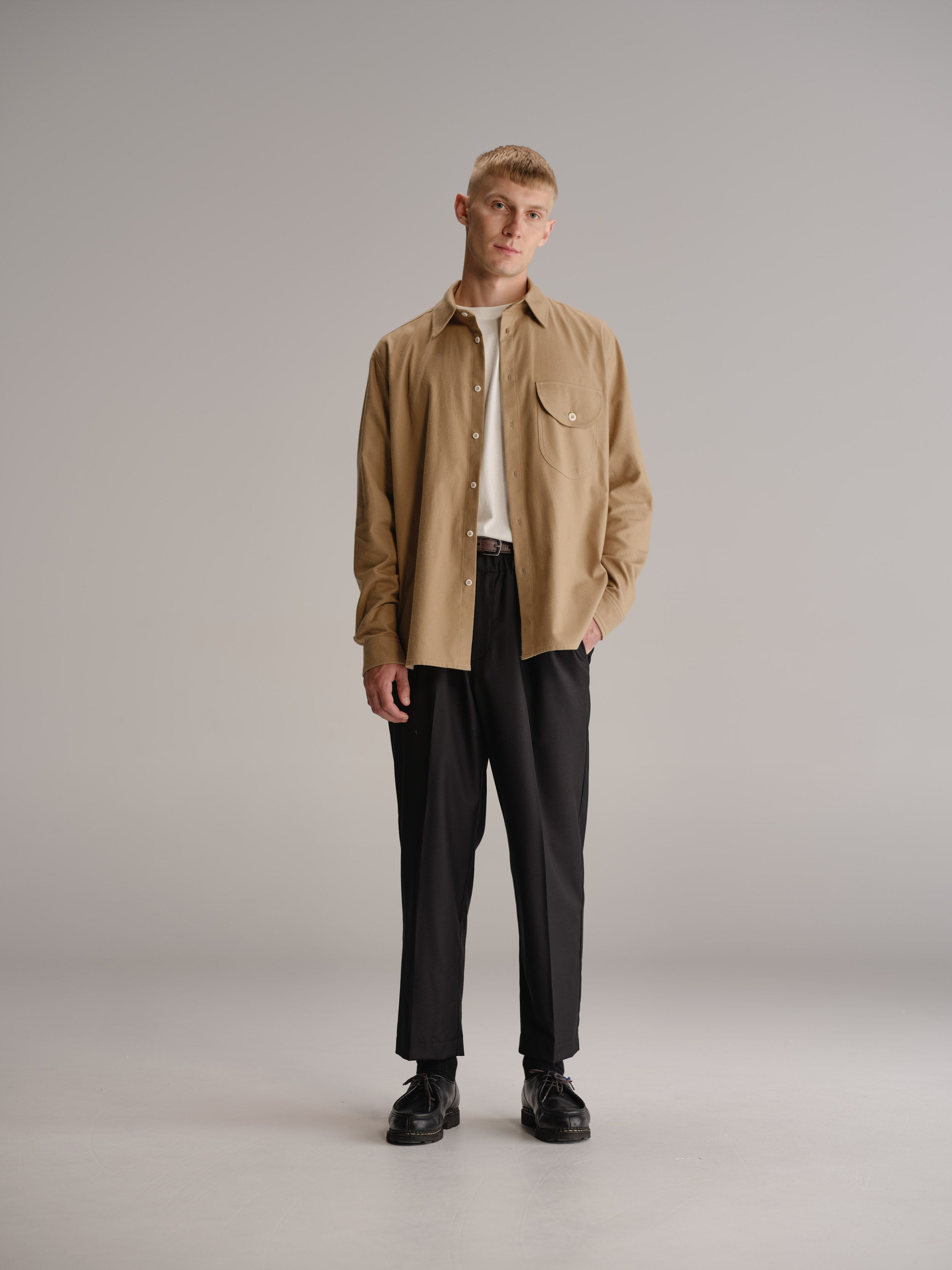 Front view of man standing in white studio wearing fawn shirt, tucked in white t-shirt and black wool trouser.