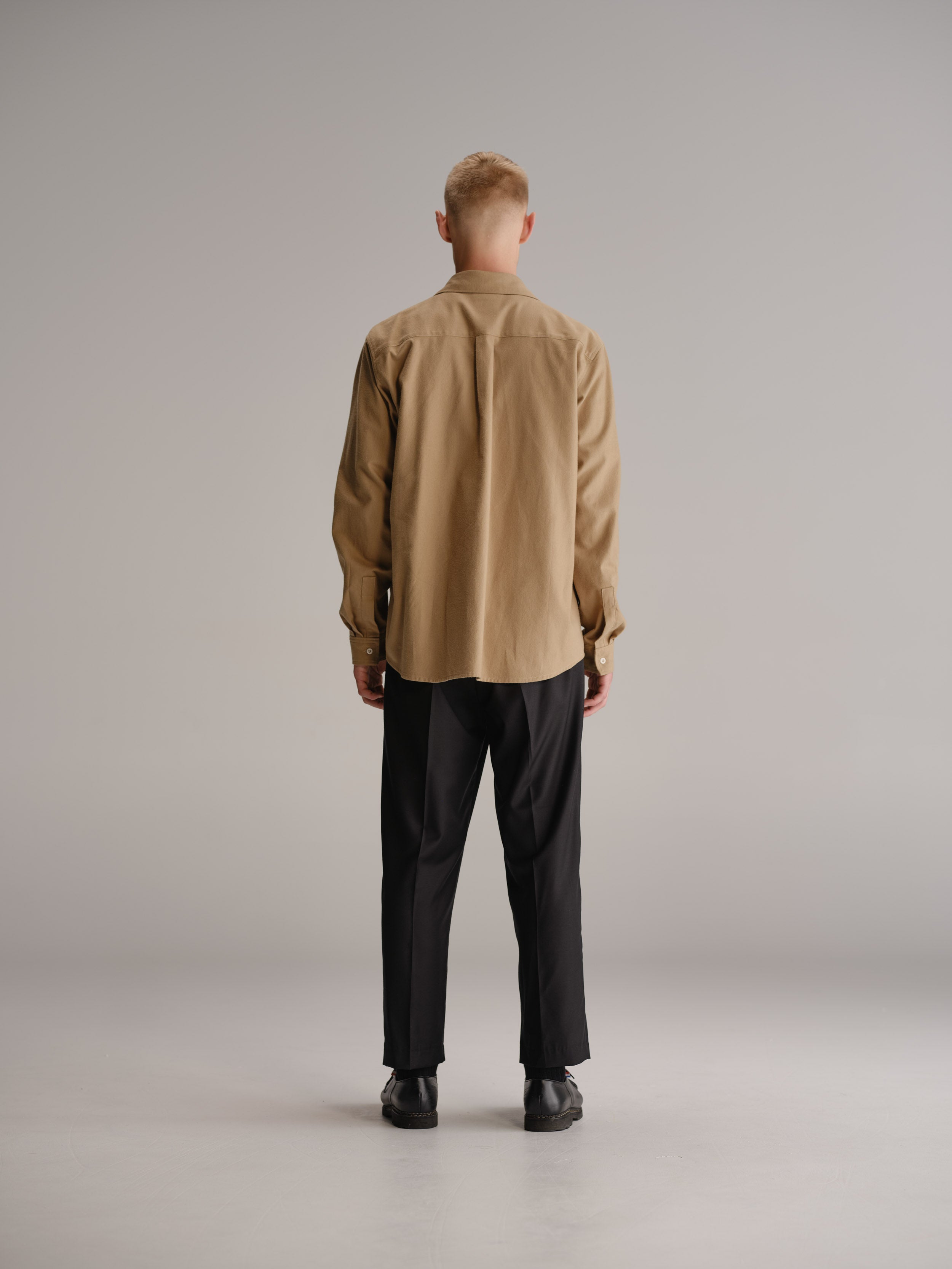 Back view of man standing in white studio wearing fawn shirt, tucked in white t-shirt and black wool trouser.