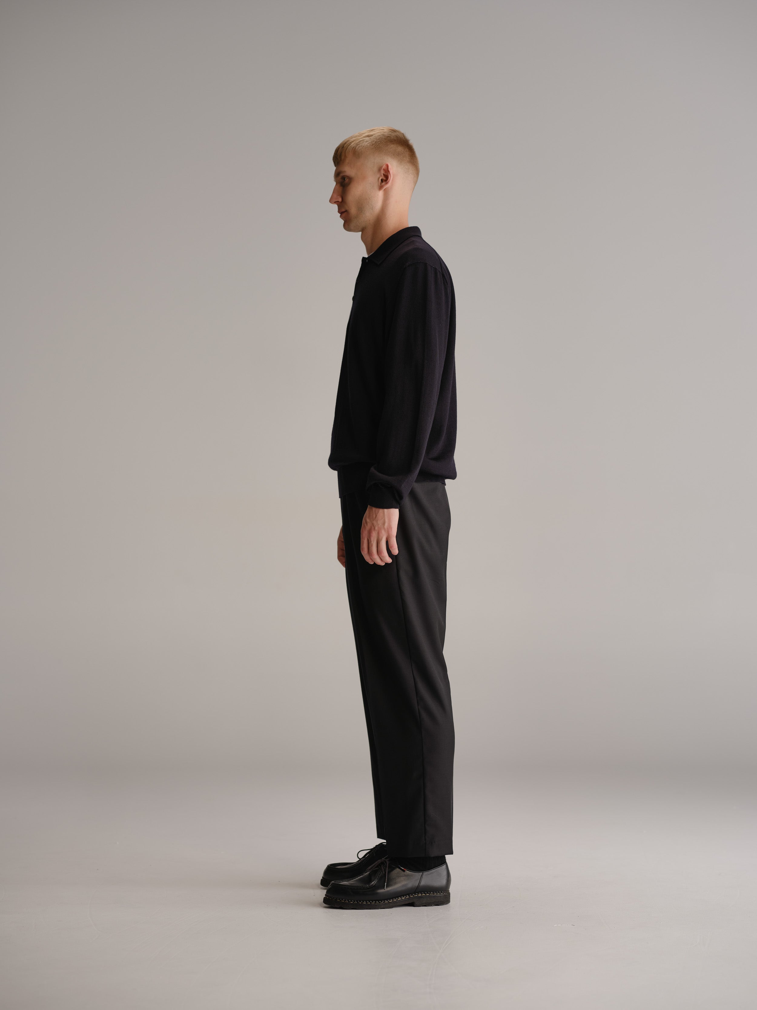 Side view of man standing in white studio wearing navy polo shirt, black wool trousers and black leather shoes.