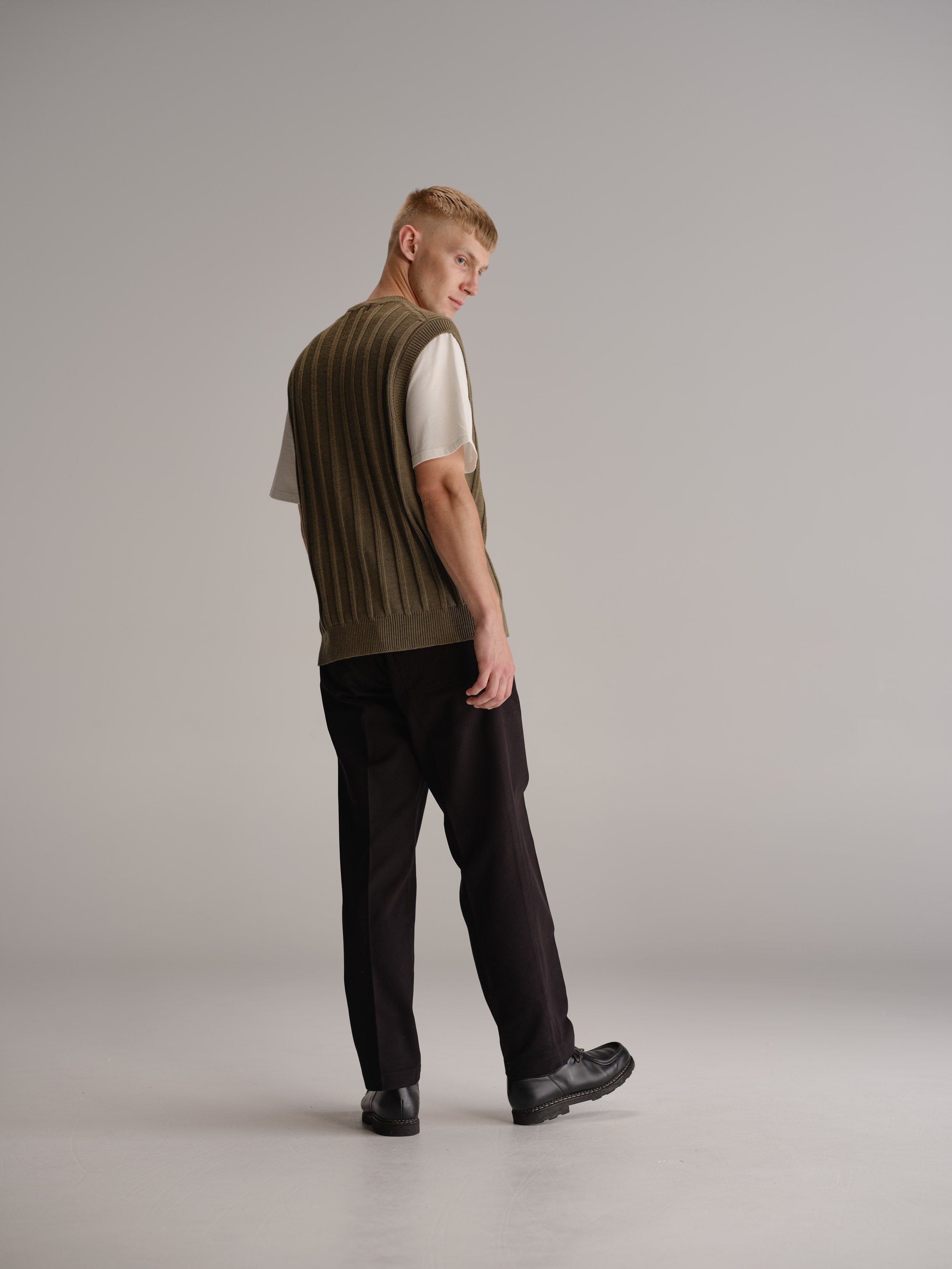 Back view of man standing in white studio wearing sage vest over a white t-shirt, black pants and black leather shoes.
