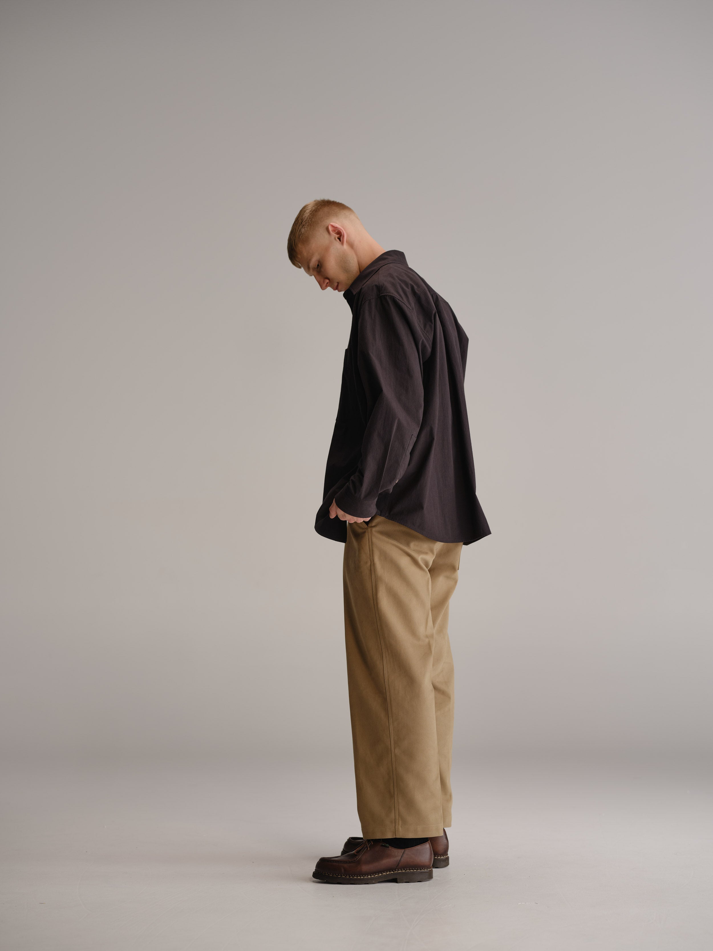 Side view of man standing in white studio wearing black shirt, tucked in black t-shirt, fawn pant and brown leather shoes.