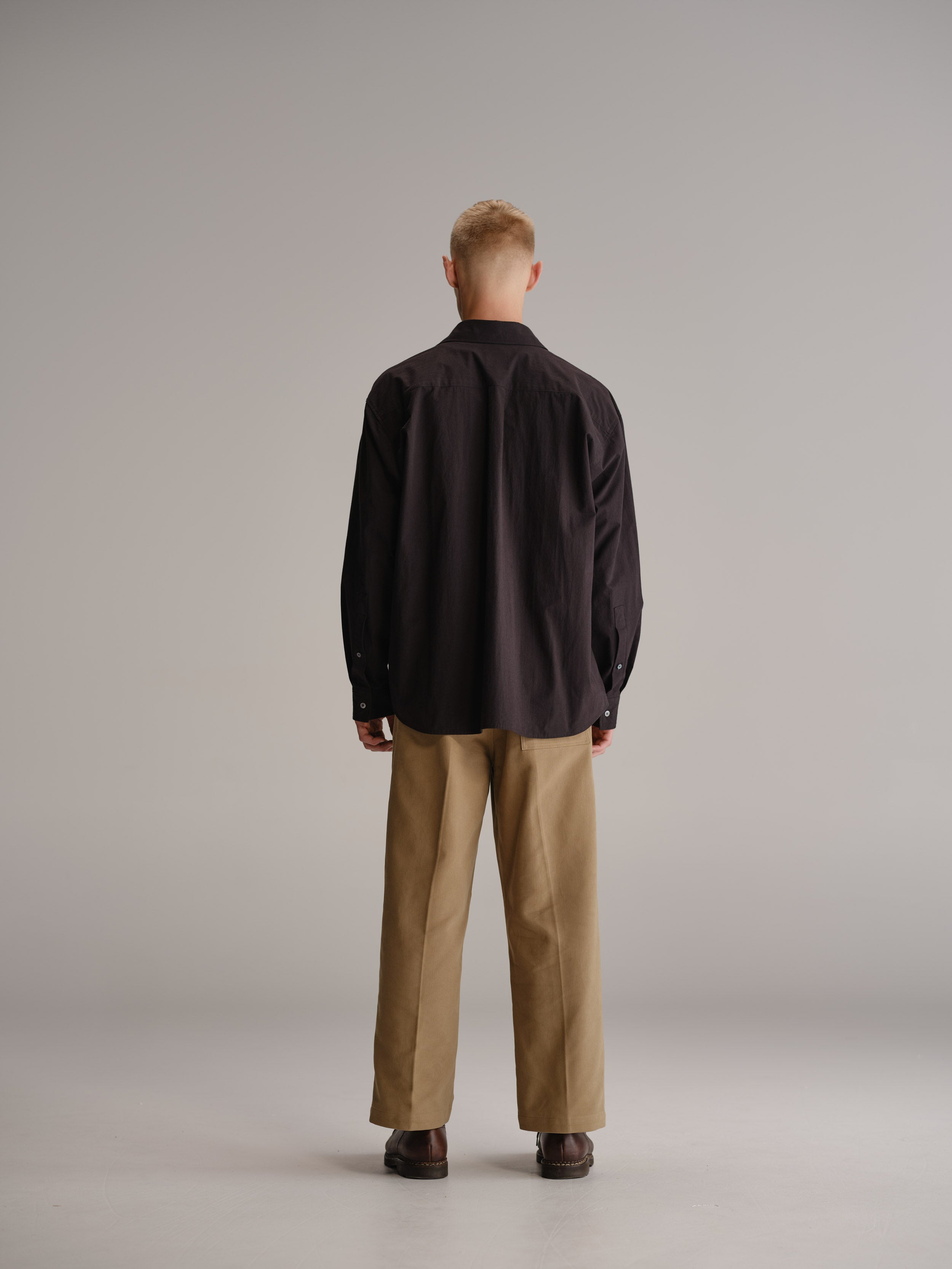 Back view of man standing in white studio with arms by side wearing black shirt, fawn pant and brown leather shoes.