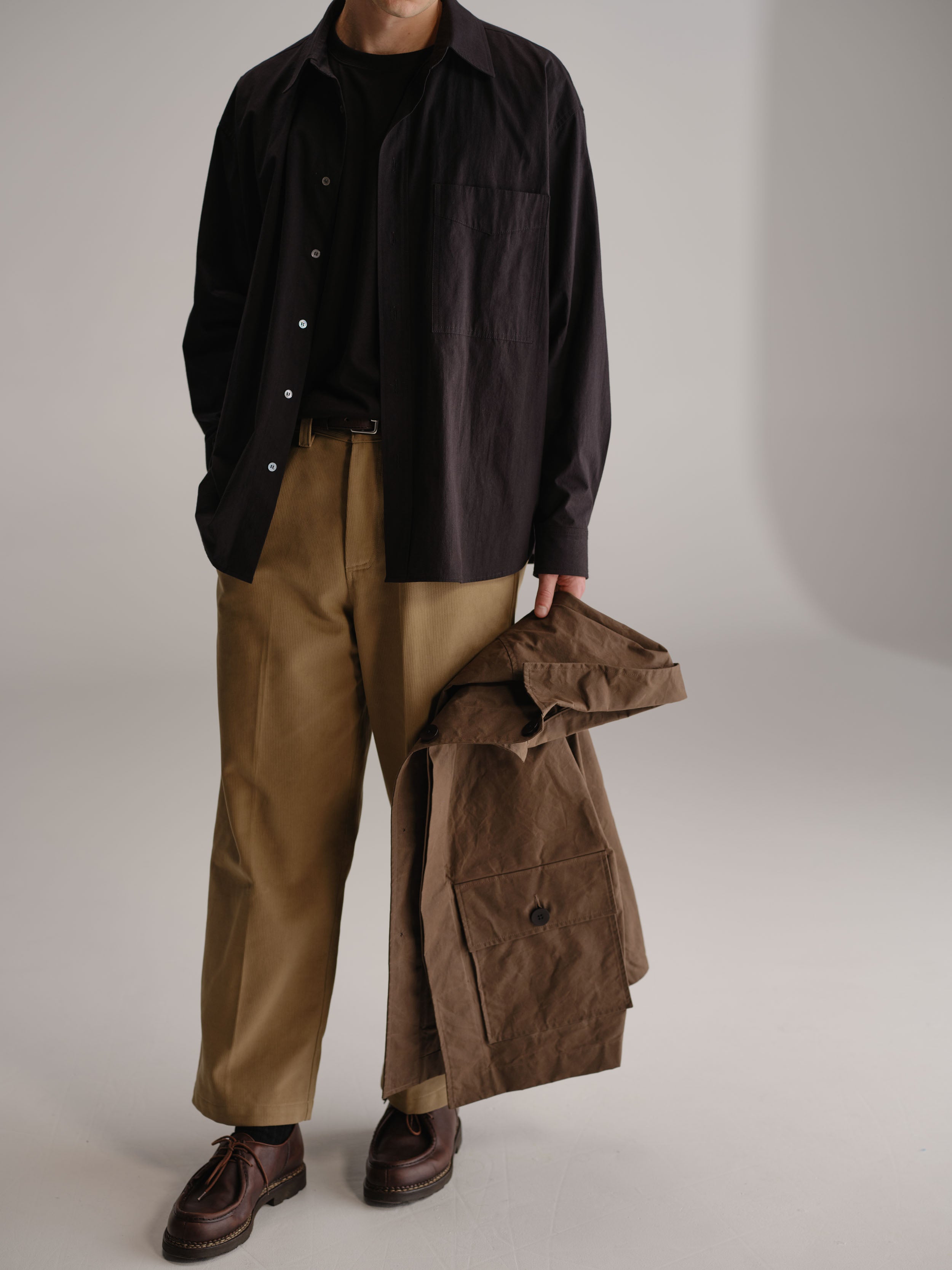 Man standing in white studio holding brown coat wearing black shirt, tucked in black t-shirt and fawn pant.