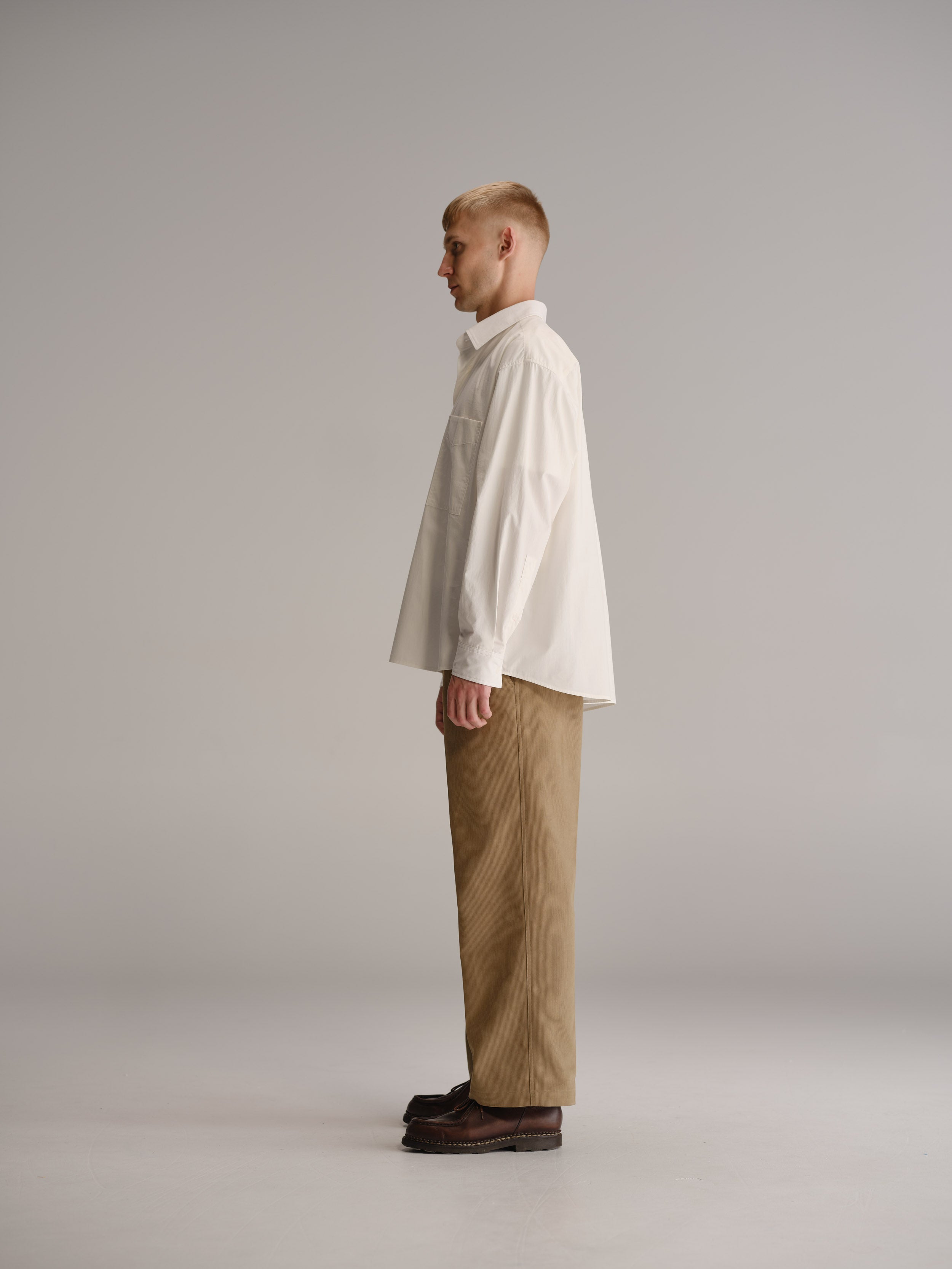 Side view of man standing in white studio wearing white shirt, fawn pants and brown leather shoes.