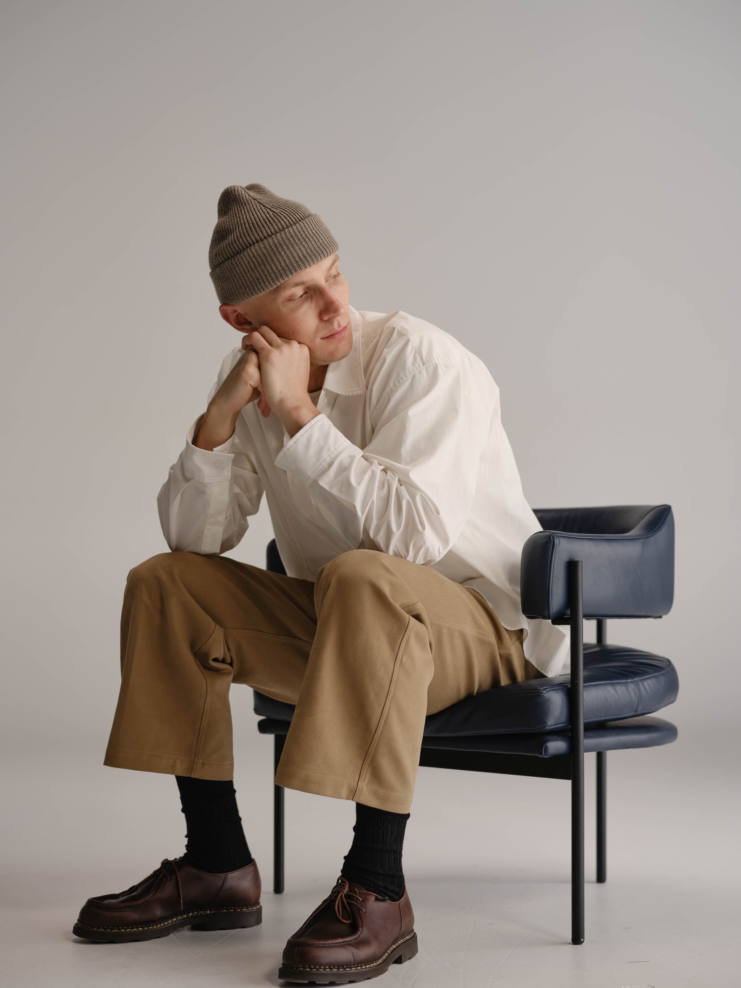 Man sitting in leather arm chair wearing wheat beanie, white shirt, fawn pants and brown leather shoes.